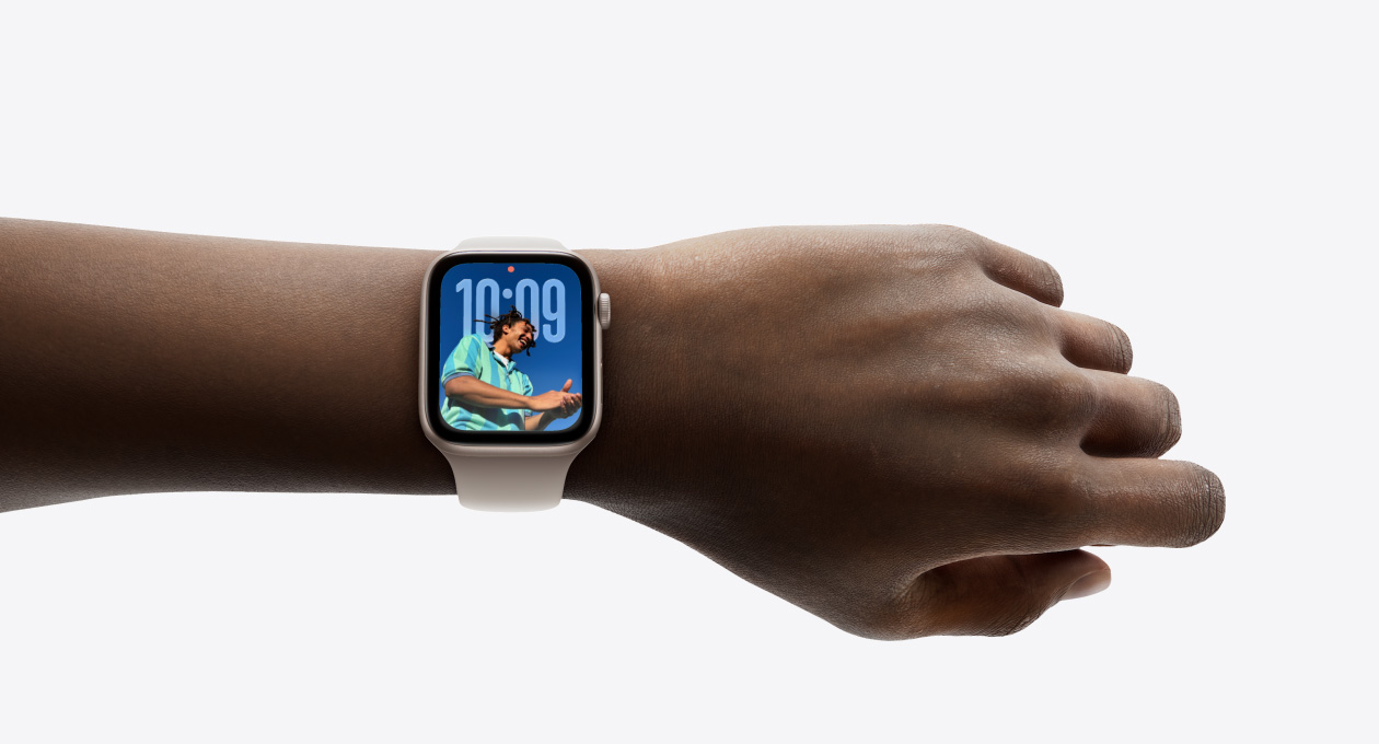 Nahaufnahme einer Apple Watch in Roségold mit Gummiarmband am Handgelenk einer Person, das Display zeigt die Uhr App im neuen Liquid Glass Design