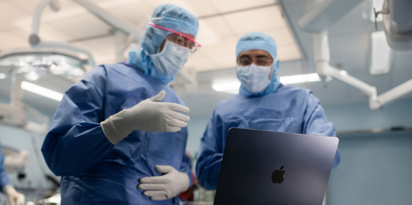 Deux chirurgiens sont dans une salle d’opération et regardent un MacBook ; l’un fait un geste de la main tandis que l’autre observe