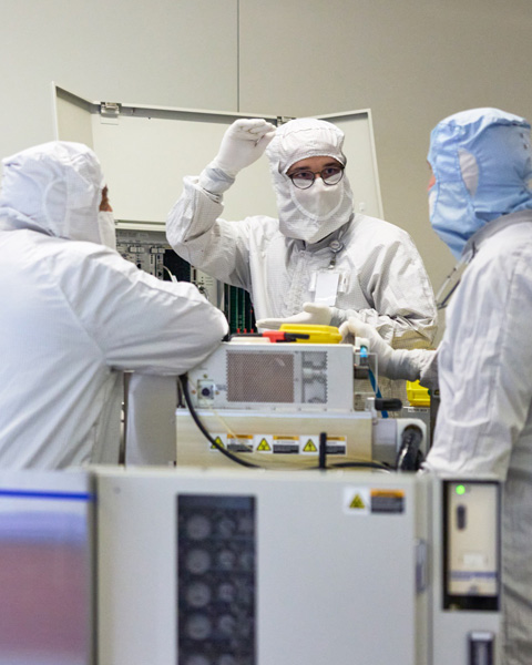 Three employees in full-body work suits stand collaborate around a silicon manufacturing machine