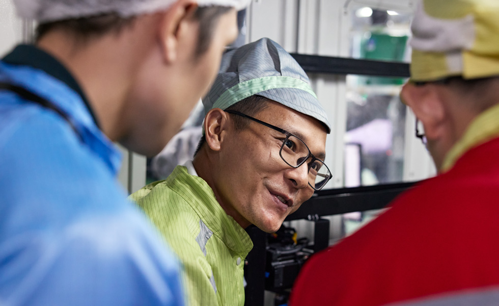 Three employees in work uniforms talk near an AirPods Pro assembly machine